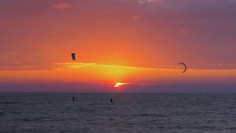 Silhouette-of-people-engaged-in-kitesurfing-in-vibrant-summer-evening-just-before-sun-goes-down,-high-contrast-beautiful-red-sunset,-Baltic-Sea-coastline,-Karosta-beach-in-Liepaja,-wide-distant-shot