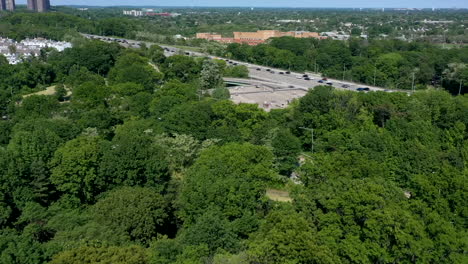 An-aerial-time-lapse-over-a-park-with-green-trees,-a-parkway-with-traffic-zooming-by-on-the-right