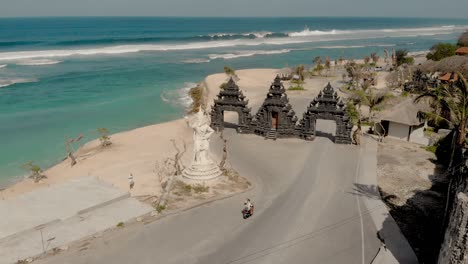 Tourist-riding-motorbikes-at-gate-to-Melasti-Beach,-Bali,-Indonesia,-aerial-view