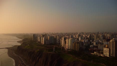 Aerial-view-overlooking-the-Costa-Verde-coast-and-the-cityscape-of-Miraflores,-sunset-in-Lima,-Peru---pan,-drone-shot