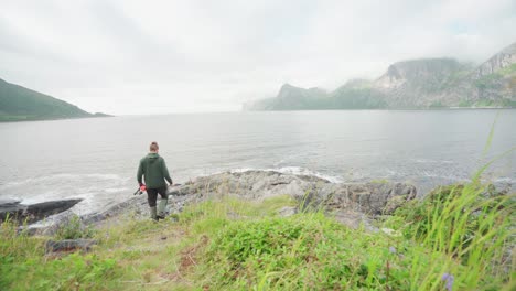 Back-View-Of-A-Man-Going-Fishing-Walking-Towards-A-Rocky-Shore-Holding-A-Fishing-Rod-In-Segla,-Senja-Island,-Norway