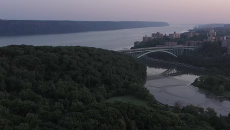 gorgeous-aerial-blue-hour-flight-over-Inwood-Hill-Park-towards-the-Henry-Hudson-Bridge-at-the-tip-of-Manhattan-New-York-City,-views-and-Spuyten-Duyvil-the-Palisades-of-New-Jersey-in-the-distance