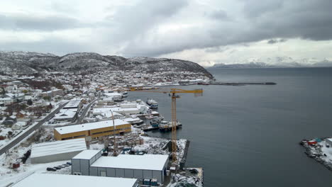 Drone-reveals-snowy-winter-landscape-of-fishing-town-in-Arctic-circle,-crane-at-harbour