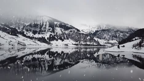 Winter-mountains-reflection-of-lake-water-during-heavy-snowfall,-aerial-drone-view