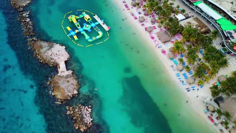 Aerial-view-dolly-in-Mambo-Beach,-inflatables-in-the-water,-Curacao,-Dutch-Caribbean-island