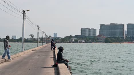 Pescador-Sentado-En-El-Muelle-Fumando-Mientras-Sostiene-Su-Caña-Y-Luego-Otro-Se-Acerca-Para-Arrojar-Algo-De-Material-De-Chumming-En-El-Agua,-Muelle-De-Pesca-De-Pattaya,-Chonburi,-Tailandia