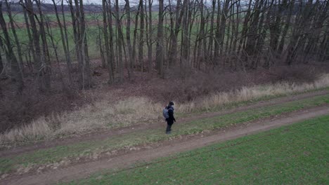 A-man-in-a-black-coat-and-hat-walking-with-a-backpack-along-a-row-of-trees-along-a-path-near-a-field-in-spring