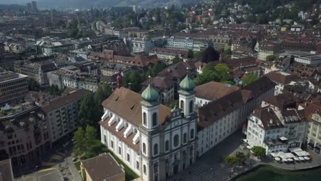 Aerial-orbit-of-baroque-Jesuit-Church-in-Lucerne-Old-Town-next-to-Reuss-River,-Chapel-Bridge-and-Water-Tower,-hills-in-background,-Switzerland