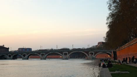 Students-enjoying-the-sunset-on-the-Garonne-riverbank-next-to-the-Pont-Neuf-bridge,-Toulouse,-France