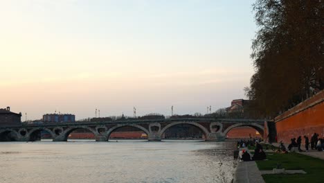 Estudiantes-Disfrutando-De-La-Puesta-De-Sol-En-La-Orilla-Del-Río-Garona-Junto-Al-Puente-Pont-Neuf,-Toulouse,-Francia