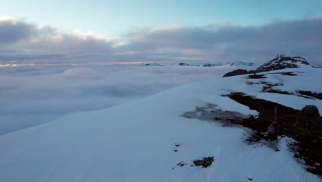 Luftüberflug-Eines-Bergsteigers,-Der-Am-Rand-Einer-Schneebedeckten-Klippe-Steht-Und-Eine-Wolkenbedeckte-Landschaft-Mit-Tiefstehender-Sonne-Freigibt