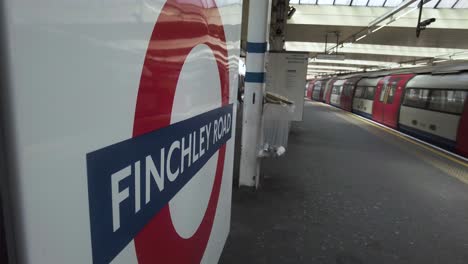 Vista-Lateral-De-Cerca-Del-Cartel-De-La-Estación-De-Roundel-De-Finchley-Road-Con-El-Tren-Jubilee-En-Dirección-Norte-Que-Llega-A-La-Plataforma-El-12-De-Mayo-De-2022