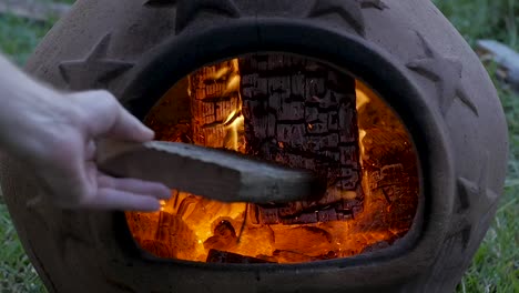 SLO-MO:-Caucasian-male-bumps-burning-embers-in-a-terra-cotta-chimenea-with-a-piece-of-wood-during-late-evening
