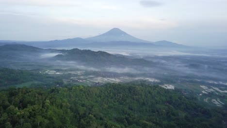 Aerial-view-over-green-forest-trees,-clouds-and-fog-in-the-valley-and-MOunt-Sumbing-in-background