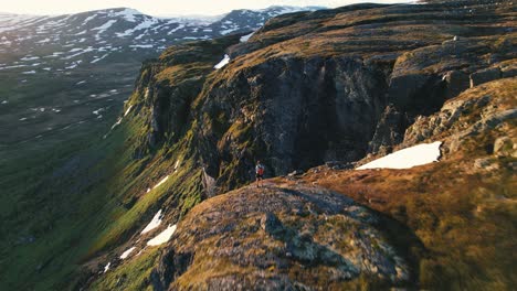 Antena-A-Lo-Largo-Del-Borde-Superior-De-La-Montaña-Iluminada-Por-El-Sol-Con-Un-Excursionista-De-Pie-En-El-Parque-Nacional-Hardangervidda,-Eidfjord,-Noruega