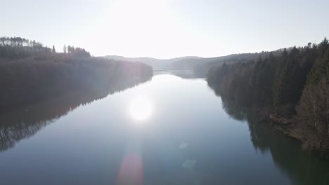 Drone-flight-towards-the-low-sun-over-a-blue-fresh-water-reservoir-within-a-brown-forest-landscape