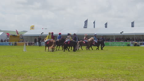 Niños-Pequeños-Vestidos-Con-Sedas-De-Jockey-Coloridas-Montando-Ponis-Dando-Vueltas-En-Preparación-Para-Una-Carrera---Cámara-Lenta