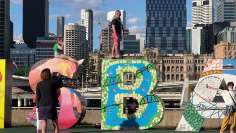 Male-tourist-standing-high-on-top-of-the-artistic-block-letter,-having-fun-taking-photos-with-the-iconic-landmark-of-Brisbane-city-on-a-sunny-day,-Queensland,-Australia,-close-up-handheld-motion-shot