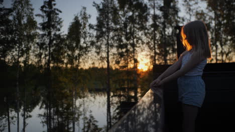 Young-Blonde-Girl-enjoys-sunset-lake-view-at-relaxing-nordic-summer-cabin,-Scandinavian-Vacation-Scene