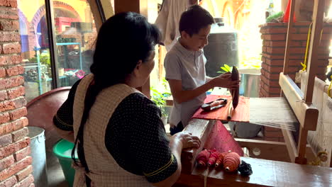 Dentro-De-La-Fabricación-Tradicional-De-Talleres-De-Alfombras-Artesanales,-Mujer-Enseñando-A-Ver-A-Un-Niño-Practicando-La-Manipulación-De-La-Máquina-Trenzadora,-Aprendiz-De-Artesano-Trabajando,-Artesanía-Artesanal-De-Telares-Manuales