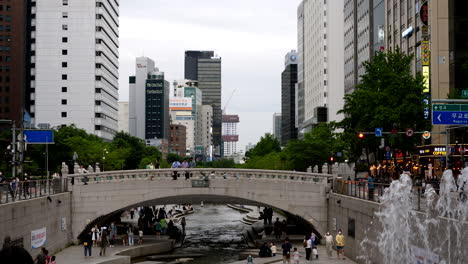 Toma-En-Cámara-Lenta-De-Personas-En-El-Puente-Gwangtong-Y-Arroyo-En-El-Centro-De-La-Ciudad-De-Seúl---área-De-Cheonggyecheon-Rodeada-De-Rascacielos