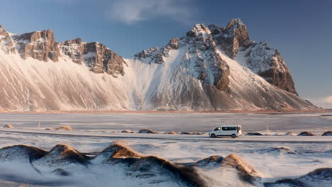 Aerial-View-Tracking-Van-Moving-along-Snowy-Road-in-Vestrahorn-Wild-Area-Iceland,-Surrounded-by-Spectacular-Winter-Landscape-and-Rocky-Mountain