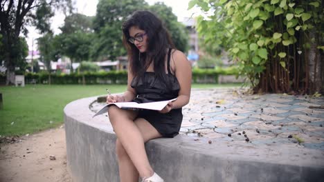 A-hot-Asian-girl-is-studying-in-the-University-College-Campus,-writing-in-a-notebook-under-a-tree