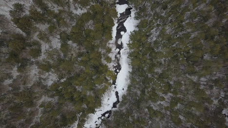 Top-down-aerial-shot-of-drone-going-up-above-snowy-river-in-winter