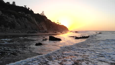 Ein-Strand-Bei-Sonnenaufgang-Mit-Meereswellen-Und-Reflexionen-Am-Ufer-Und-Klippen-In-Silhouette-Entlang-Der-Luftdrohne-An-Der-Küste-Von-Santa-Barbara,-Kalifornien