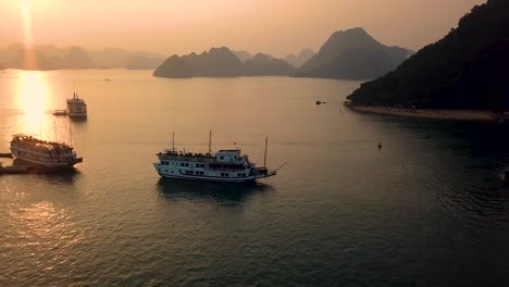 Aerial-shot-moving-slowly-towards-ships-in-Ha-Long-Bay-with-beautiful-sunset-and-island-cliffs-in-background