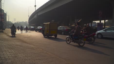 Traffic-on-the-road-under-the-fly-over,-passing-motor-bikes-and-cars,-Buses,-rickshaw`s,-Billboards,-buildings,-electric-polls-and-street-lights,-A-biker-without-helmet-coming-from-wrong-side