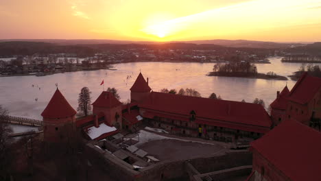 AERIAL:-Wide-Shot-of-Trakai-Island-Castle-with-Orange-Sunset-and-Frozen-Lake-in-the-Background