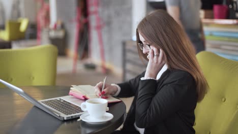 Mujer-Tomando-Notas-Mientras-Habla-Por-Teléfono-Frente-A-Una-Laptop-En-Una-Cafetería