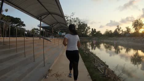 A-slowmotion-shot-of-a-young-thai-girl-jogging-on-a-pavement-near-a-small-lake-during-sunset,-Thailand