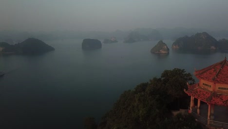 Aerial-shot-slowly-moving-over-red-temple-on-top-of-cliff-to-reveal-Ha-Long-Bay-at-dusk-as-sun-sets