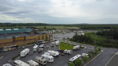 percent-hyperlapse-aerial-flyover-of-a-Cabela's-parking-lot-in-Anchorage,-Alaska-showing-many-recreational-vehicles-parked-with-cars-moving,-people-walking,-and-mountains-in-the-background