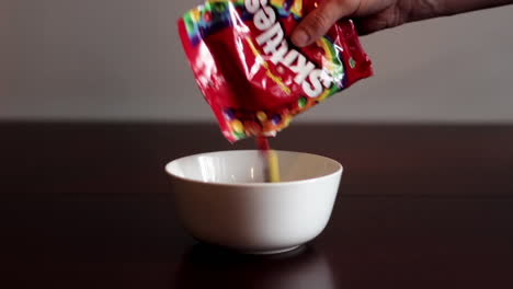 Close-Up-Hand-Pouring-Bag-Of-Skittles-Candy-Into-White-Bowl-On-Table