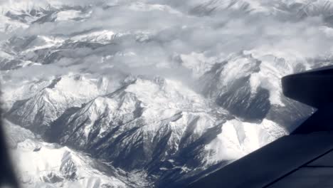 Flying-over-snow-capped-mountains-and-clouds-in-Europe-looking-out-jet-plane-window-mountians-revealed-beneath-wing