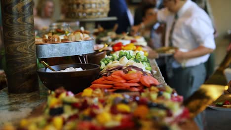 Smooth-shot-of-a-line-of-people-at-a-crowded-event-going-through-a-food-line-and-grabbing-a-variety-of-food-including-meats,-cheeses,-bread,-fruits,-and-vegetables