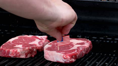 Zwei-Saftige-Rib-Eye-Steaks-Sitzen-Auf-Dem-Grill-Und-Kochen-Eine-Hand-Kommt-Herein-Und-Steckt-Mit-Einem-Zahnstocher-Eine-Winzige-Amerikanische-Flagge-In-Eines-Von-Ihnen