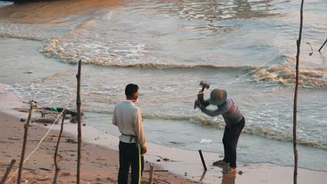 Mittlere-Außenaufnahme-Eines-Mannes,-Der-Mit-Einem-Schweren-Hammer-Einen-Pfosten-In-Den-Sand-Hämmert,-Und-Seines-Freundes-Am-Ufer-Mit-Wellen-Im-Wasser-Hinter-Ihm-Im-See-Bei-Tag