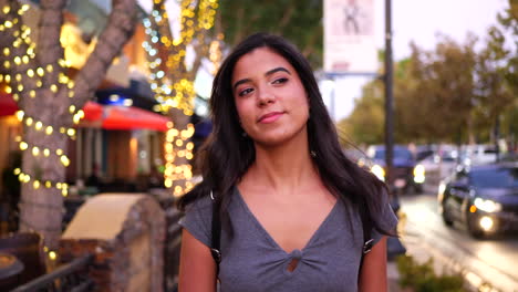 An-attractive-young-hispanic-woman-walking-a-downtown-urban-city-street-in-California-with-restaurants,-shops-and-retail-stores-with-lights-SLOW-MOTION