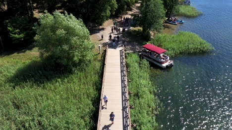 AERIAL:-Reveal-Shot-of-Long-Wooden-Bridge-and-People-Walking-on-it-on-a-Sunny-and-Bright-Day-in-Trakai-with-Green-Color-Lake
