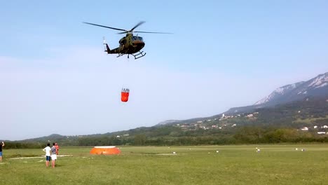 Helicopter-with-bucket-taking-water-to-fight-a-forest-fire,-firemen-with-trucks-provide-ground-support