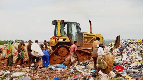 Bulldozer-plows-piles-of-garbage-at-a-dump,-poor-people-follow-the-bulldozer-and-sift-through-the-garbage