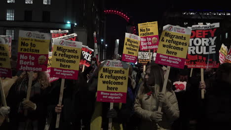 Antirassistische-Demonstranten-Stehen-Nachts-Auf-Whitehall-Und-Halten-Während-Eines-Protests-Gegen-Premierminister-Boris-Johnson-Vor-Einem-Beleuchteten-London-Eye-Plakate-Mit-Der-Aufschrift-„Stand-Up-To-Rassism“-Und-„Socialist-Worker“.