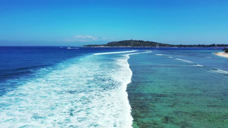 Beautiful-white-waves-of-blue-ocean-splashing-over-shallow-calm-lagoon-with-coral-reefs,-bright-clear-sky-in-Bali