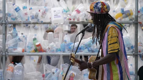 Artista-Callejero-Cantando-En-Una-Actuación-En-El-Parque,-Barcelona,-Frente-A-Una-Obra-De-Arte-En-La-Pared-De-Una-Botella-De-Plástico-Contra-La-Contaminación-Plástica.