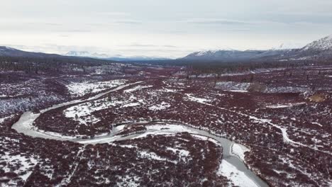 Picturesque-scenic-autumn-flight-above-frozen-O'Donnel-river-and-red-color-woods-forest-towards-mountain-range-in-background-on-bright-cloudy-day,-British-Columbia,-overhead-aerial-approach
