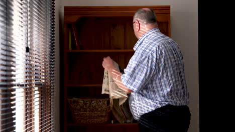 A-middle-aged-man-picks-up-a-newspaper-and-reads-then-folds-and-walks-away-while-the-sunlight-comes-through-the-Venetian-blinds